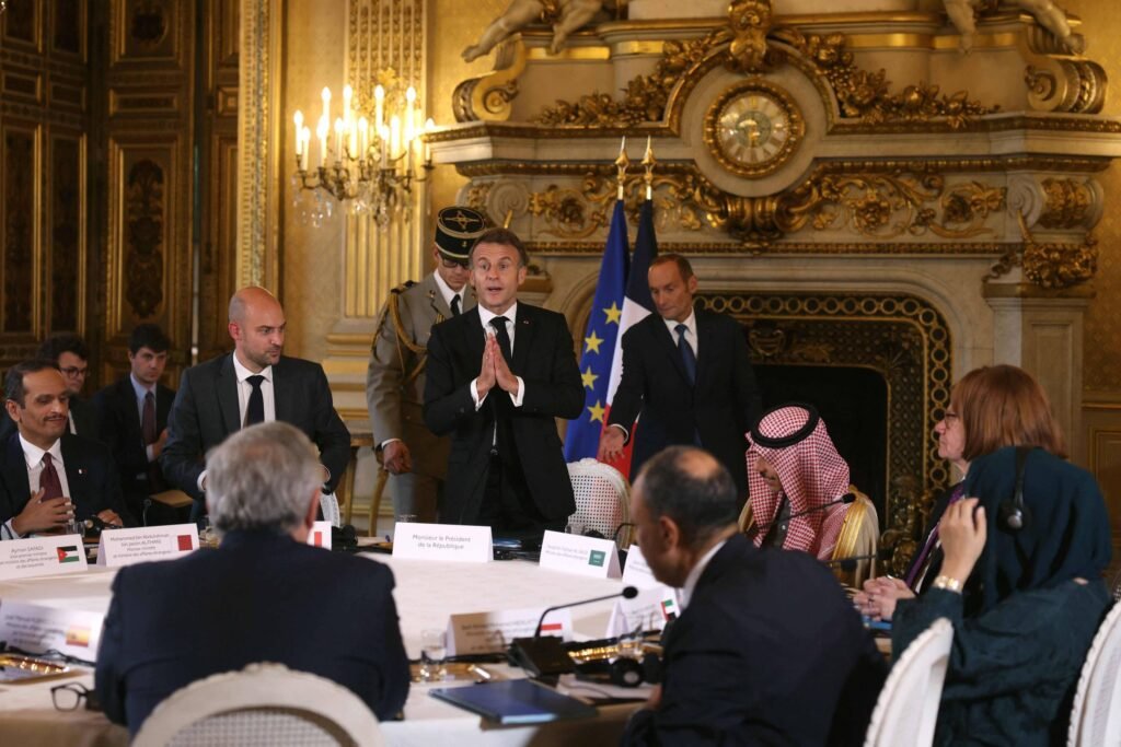 Homem em terno preto fala em pé diante de mesa redonda com participantes sentados, incluindo homens com trajes tradicionais árabes. Ambiente interno luxuoso com lareira decorada, candelabros e painéis dourados. Bandeiras da União Europeia e França ao fundo.