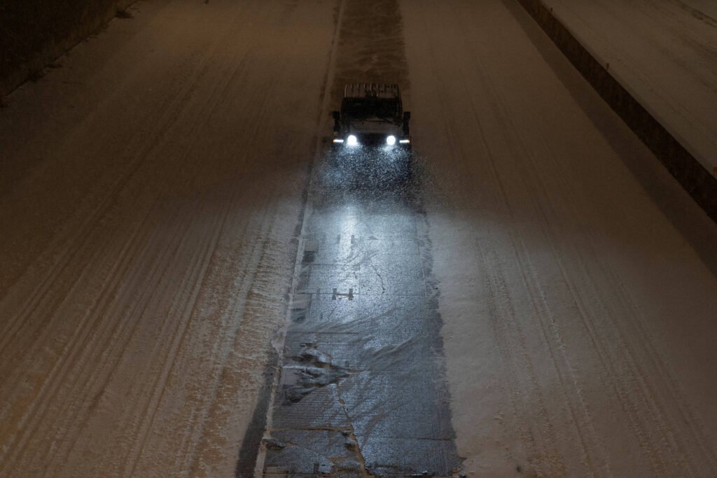 Veículo equipado com luzes frontais acesas limpa uma faixa de neve acumulada em estrada larga durante a noite, deixando o asfalto visível.