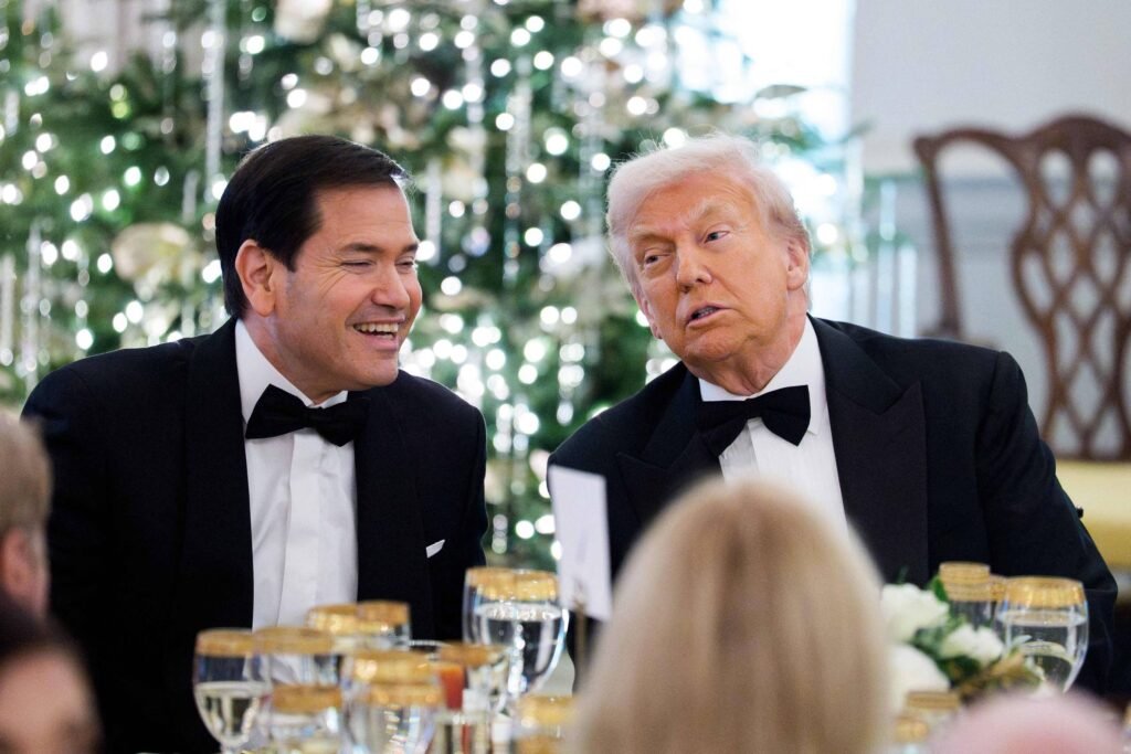 Dois homens de terno preto e gravata borboleta conversam sentados à mesa decorada com taças douradas e arranjo floral. Ao fundo, árvore de Natal iluminada e cadeira de madeira.