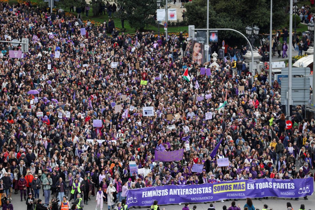 Milhares de pessoas participam de manifestação feminista em rua movimentada, segurando cartazes e faixas roxas. Faixa principal na frente diz: