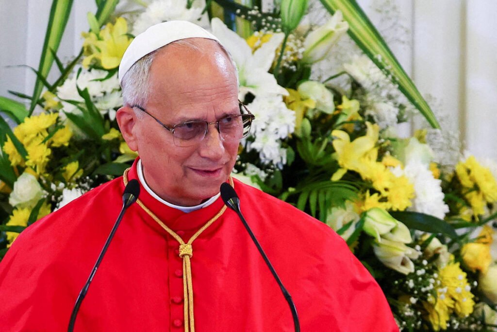 Homem idoso com solidéu branco e vestes vermelhas fala em microfones diante de arranjo floral com flores brancas e amarelas.