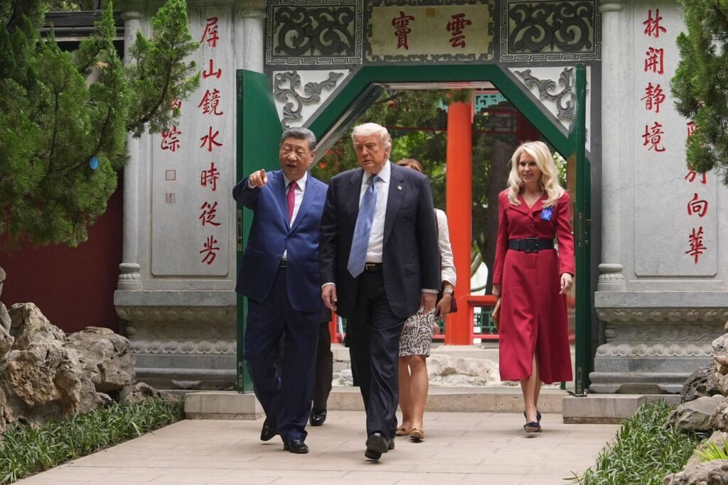 Dois líderes caminham lado a lado em um caminho de pedra dentro de um jardim tradicional chinês, com uma mulher atrás deles. O homem à esquerda veste terno azul e gravata vermelha, apontando para algo à frente. O homem à direita usa terno escuro e gravata azul. A mulher veste vestido vermelho com cinto preto. Ao fundo, há um portal decorado com caracteres chineses vermelhos e verdes.
