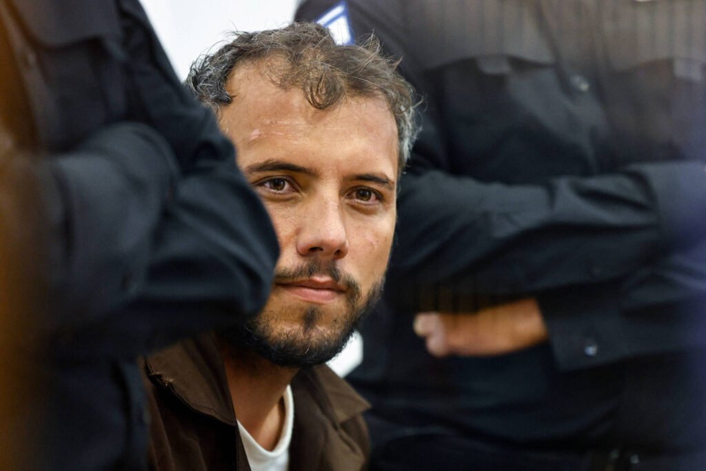 Homem de cabelos curtos e barba curta olha diretamente para a câmera, cercado por policiais vestidos com uniformes pretos. O foco está no rosto do homem, com os policiais parcialmente visíveis ao redor, em ambiente interno.