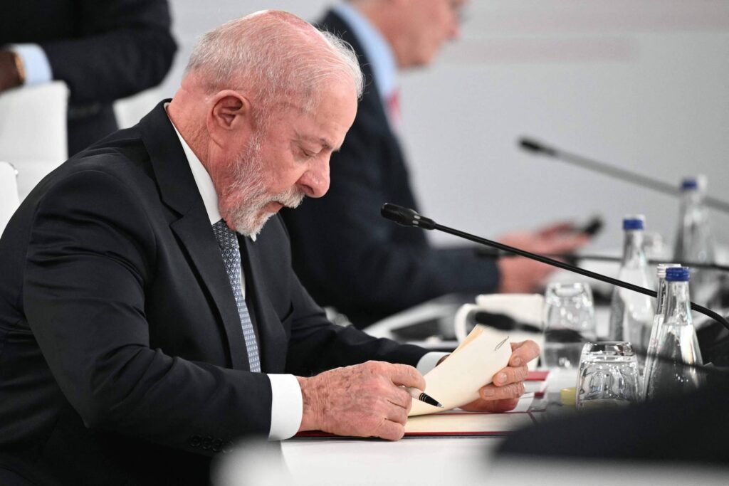 Homem idoso de terno escuro e gravata está sentado à mesa, lendo atentamente um documento. Há microfone, copos de vidro e garrafas de água sobre a mesa. Ao fundo, outra pessoa está desfocada, também sentada.